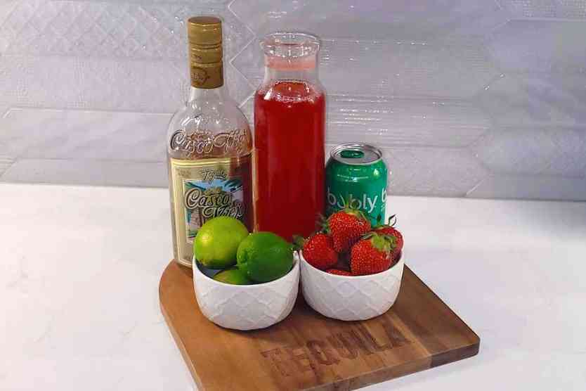 Cutting board with strawberry simple syrup, strawberries, limes, soda water and Tequila.